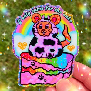 May include: A holographic sticker featuring a cartoon creature with cow-like spots and small horns sitting on a multi-layered cake. The text "I only came for the cake" is arched above the creature, set against a rainbow and two orange hearts.