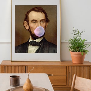 Pode incluir: Um retrato emoldurado de um homem com barba soprando uma bolha de chiclete rosa. O retrato est&aacute; sobre um fundo marrom e tem uma moldura dourada.
