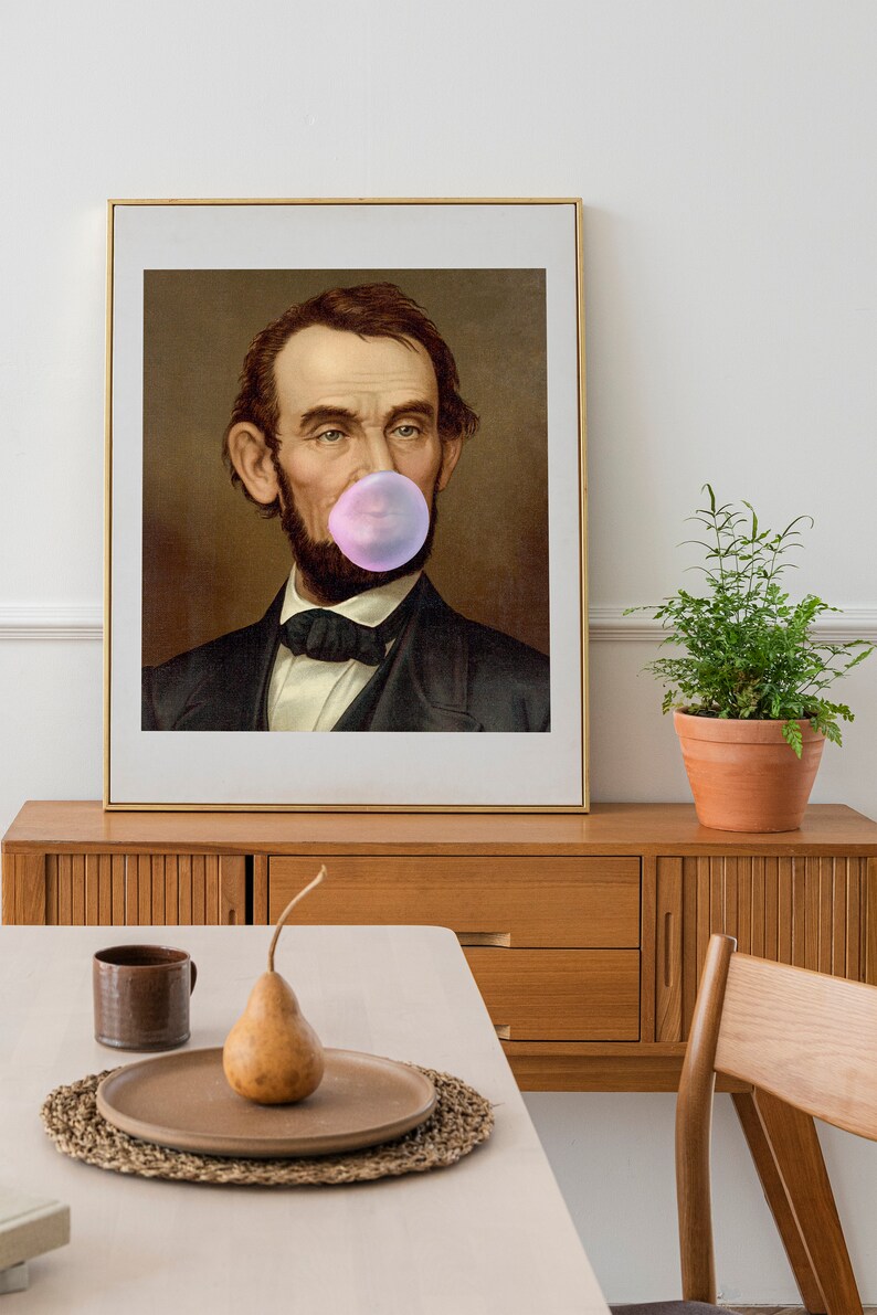 Pode incluir: Um retrato emoldurado de um homem com barba soprando uma bolha de chiclete rosa. O retrato est&aacute; sobre um fundo marrom e tem uma moldura dourada.