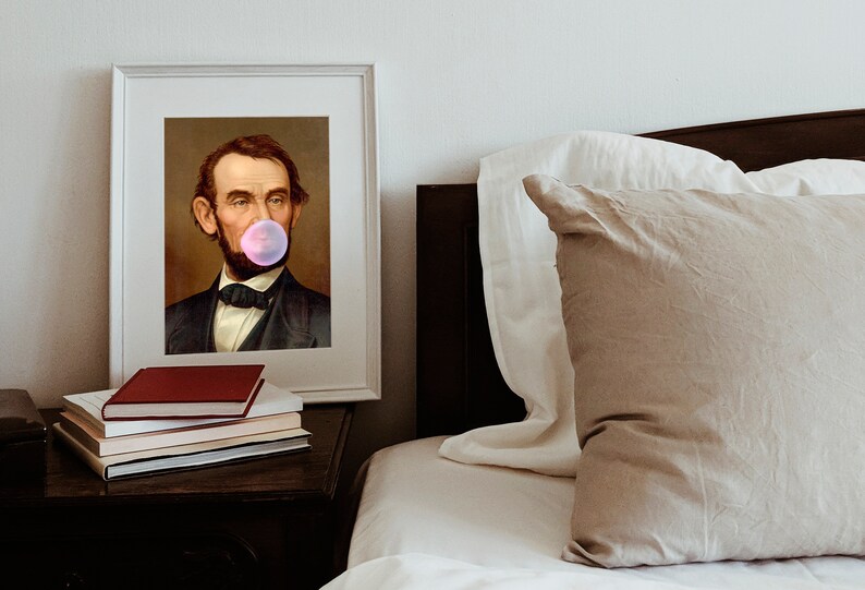 Pode incluir: Um retrato emoldurado de um homem com barba soprando uma bolha de chiclete rosa. O retrato est&aacute; pendurado em uma parede acima de uma mesa de cabeceira com uma pilha de livros.