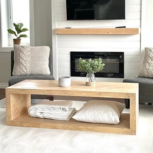 Puede incluir: Una mesa de centro de madera clara con un estante abierto rectangular. La mesa tiene una manta blanca y una almohada blanca en el estante. La mesa está delante de una chimenea con una pantalla negra y una repisa blanca.