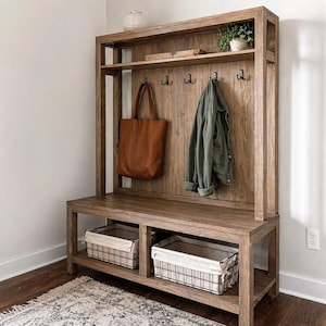 May include: A wooden entryway bench with a built-in shelf and coat rack. A brown leather tote bag and a green jacket hang from the hooks. Two wire baskets with fabric liners sit below the bench. A small plant and decorative items are on the top shelf.