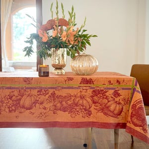 Könnte beinhalten: Eine rot-orange Tischdecke mit einem Herbst-Ernte-Design mit Kürbissen, Trauben und Mais. Die Tischdecke liegt auf einem Holztisch mit einer Blumenvase, einer Kerze und einem dekorativen Kürbis.