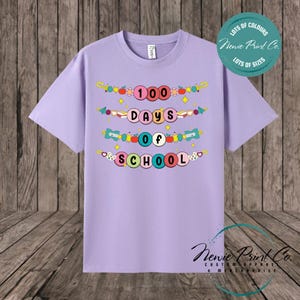 May include: A lilac t-shirt with the words "100 Days of School" in colourful bubble letters. The shirt also features decorative elements like apples and stars. The shirt has the text "Lots of Colours" and "Lots of Sizes" on it.