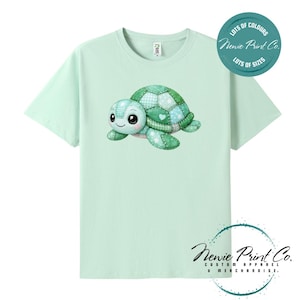 May include: A light mint green t-shirt featuring a central graphic of a cartoon turtle with a patchwork-style shell in shades of green and white. The top right corner includes a circular teal badge with the text "LOTS OF COLOURS Newie Print Co. LOTS OF SIZES".