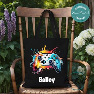 May include: Black tote bag with a colourful paint splatter design featuring a video game controller. The bag has the name "Bailey" printed in white below the design.