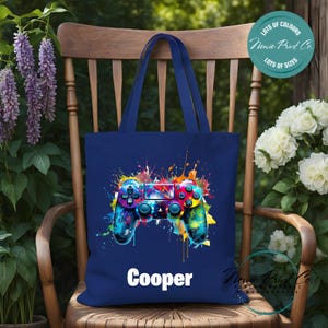 May include: A navy blue tote bag featuring a colourful paint splatter design of a video game controller. The bag has a white personalised name "Cooper" printed on the front.