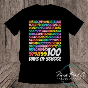 May include: Black t-shirt with a colourful graphic design of numbers counting up to 100. The text "Days of School" is below the numbers. The text "Newie Print Co. Custom Apparel & Merchandise" is at the bottom of the shirt.