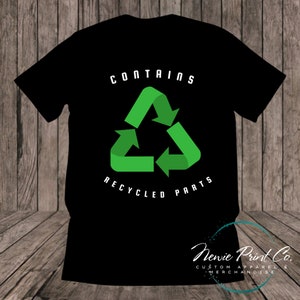 May include: Black t-shirt featuring a green recycling symbol and the text "CONTAINS RECYCLED PARTS". The bottom of the shirt has the text "Newie Print Co. CUSTOM APPAREL & MERCHANDISE".