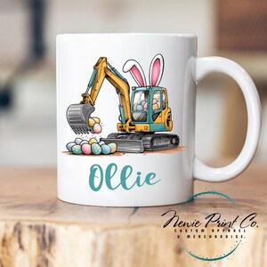 May include: A white ceramic mug featuring a cartoon yellow and blue excavator digging up Easter eggs. The excavator has bunny ears and the mug is personalised with the name "Ollie".