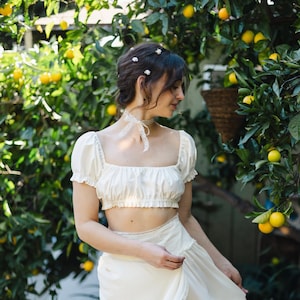 May include: A woman wearing a white crop top and a white skirt with lace trim stands in front of a lemon tree. She has her hair pulled back and is wearing small white flowers in her hair.