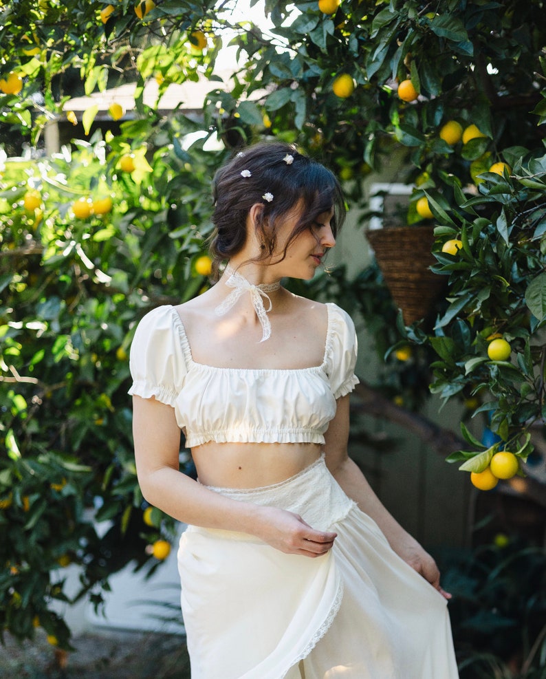 May include: A woman wearing a white crop top and a white skirt with lace trim stands in front of a lemon tree. She has her hair pulled back and is wearing small white flowers in her hair.