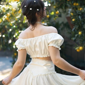 May include: A woman wearing a white off-the-shoulder crop top with a white skirt. She has her hair styled in a bouffant and is wearing small white flowers in her hair.
