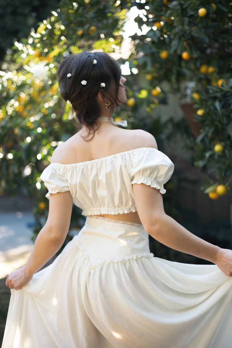 May include: A woman wearing a white off-the-shoulder crop top with a white skirt. She has her hair styled in a bouffant and is wearing small white flowers in her hair.