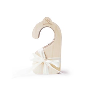 Pode incluir: Um conjunto de cabides de madeira com o texto "NB newborn" no topo. Os cabides são amarrados com uma fita branca.