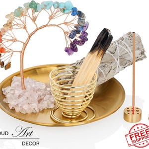Puede incluir: Una bandeja dorada de latón contiene un árbol de alambre adornado con piedras preciosas de colores, un porta incienso dorado, un manojo de salvia blanca y varillas de incienso. La bandeja también presenta un lecho de cristales de cuarzo transparente. Incluye el texto "CLOUD Art DECOR".