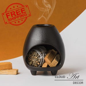 Peut inclure: Brûle-encens en céramique noire avec trois pieds. Le brûleur a une ouverture ronde à l'avant et est rempli d'herbes séchées et de copeaux de bois. Le brûleur est sur une surface blanche avec un fond brun.