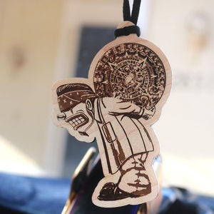 Puede incluir: Ambientador de coche de madera con un personaje de dibujos animados que lleva un gran objeto circular grabado. El personaje lleva un pañuelo y tiene una expresión seria. Cuelga de un cordón negro.