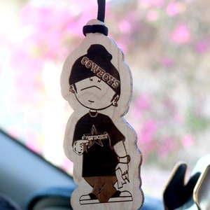 May include: Wooden car air freshener with a cartoon character wearing a black hat with the word "COWBOYS" on it. The character is wearing a black shirt with a star and the word "COWBOYS" on it.
