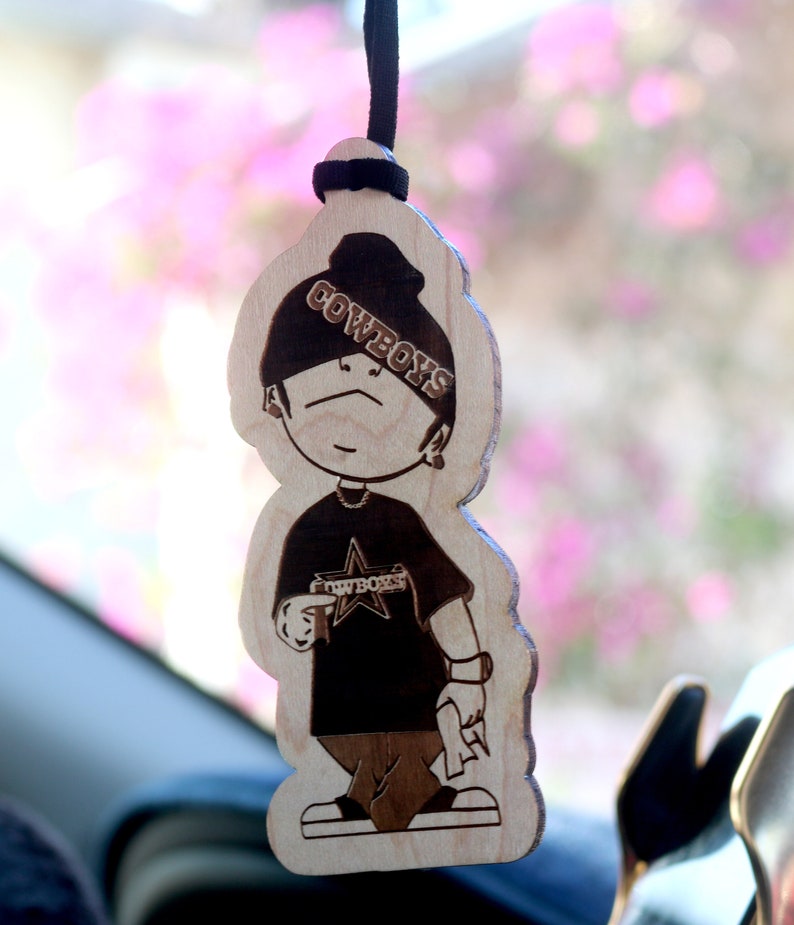 May include: Wooden car air freshener with a cartoon character wearing a black hat with the word "COWBOYS" on it. The character is wearing a black shirt with a star and the word "COWBOYS" on it.