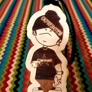 May include: A wooden cutout of a cartoon character wearing a black beanie with the word "COWBOYS" written on it. The character is wearing a black t-shirt with a star and the word "COWBOYS" written on it. The character is holding a gun in one hand and a rolled-up piece of paper in the other hand.