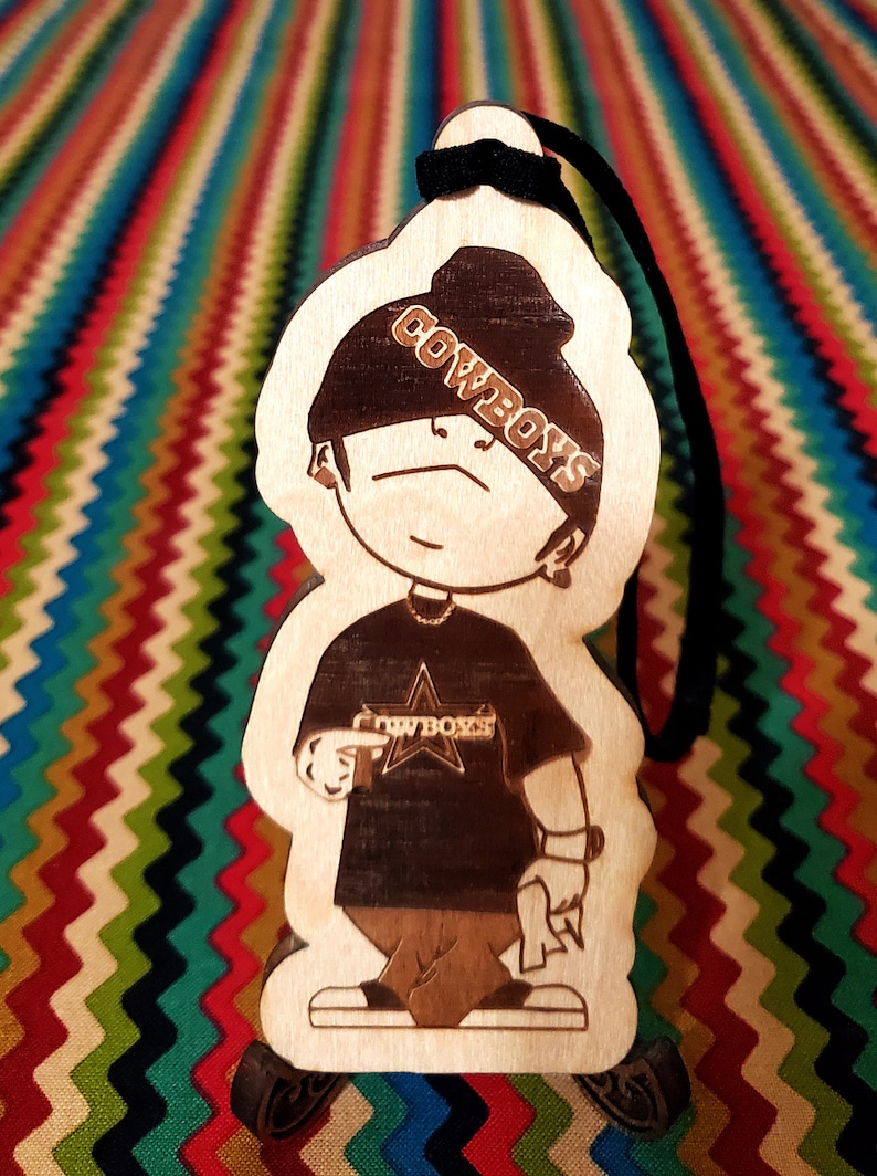 May include: A wooden cutout of a cartoon character wearing a black beanie with the word "COWBOYS" written on it. The character is wearing a black t-shirt with a star and the word "COWBOYS" written on it. The character is holding a gun in one hand and a rolled-up piece of paper in the other hand.