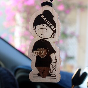 May include: A wooden car air freshener shaped like a cartoon figure wearing a black beanie with the word "RAIDERS" written on it. The figure is wearing a black shirt with a white "RAIDERS" logo and black pants.