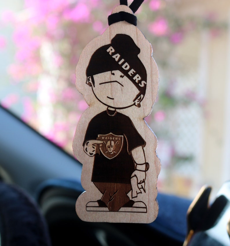 May include: A wooden car air freshener shaped like a cartoon figure wearing a black beanie with the word "RAIDERS" written on it. The figure is wearing a black shirt with a white "RAIDERS" logo and black pants.