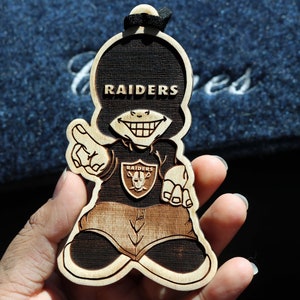 May include: A wooden ornament shaped like a cartoon character wearing a black shirt with the word "RAIDERS" on it. The character is smiling with a mischievous grin and has a Raiders logo on the front of the shirt.