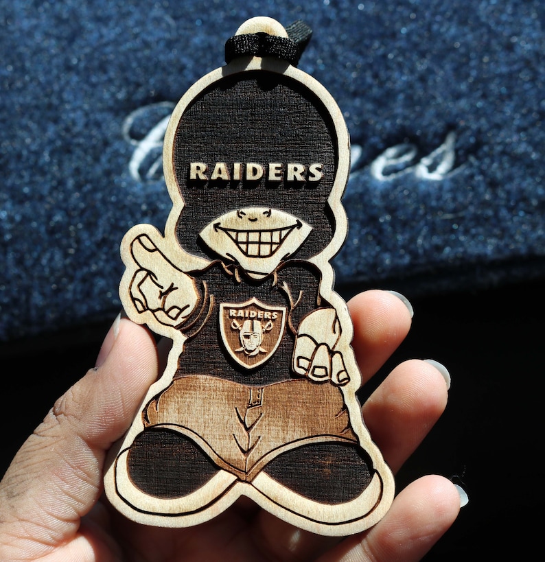 May include: A wooden ornament shaped like a cartoon character wearing a black shirt with the word "RAIDERS" on it. The character is smiling with a mischievous grin and has a Raiders logo on the front of the shirt.