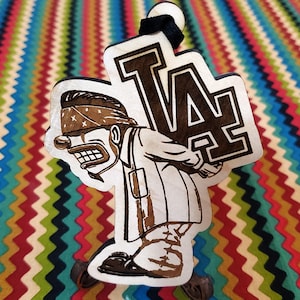 May include: A wooden ornament featuring a cartoon character wearing a bandana and carrying a large "LA" sign. The ornament is painted in brown and black.