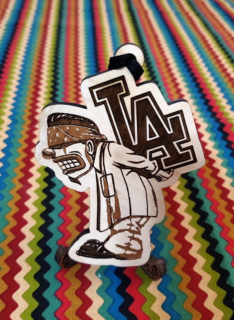 May include: A wooden ornament featuring a cartoon character wearing a bandana and carrying a large "LA" sign. The ornament is painted in brown and black.