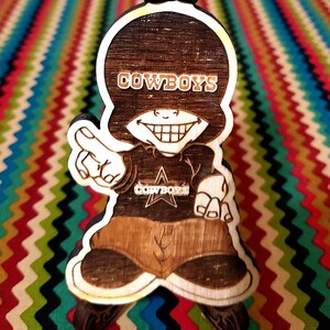 May include: A wooden figurine of a cartoon character wearing a hat and a shirt with the word "COWBOYS" on it. The character is smiling and has a star on its shirt.