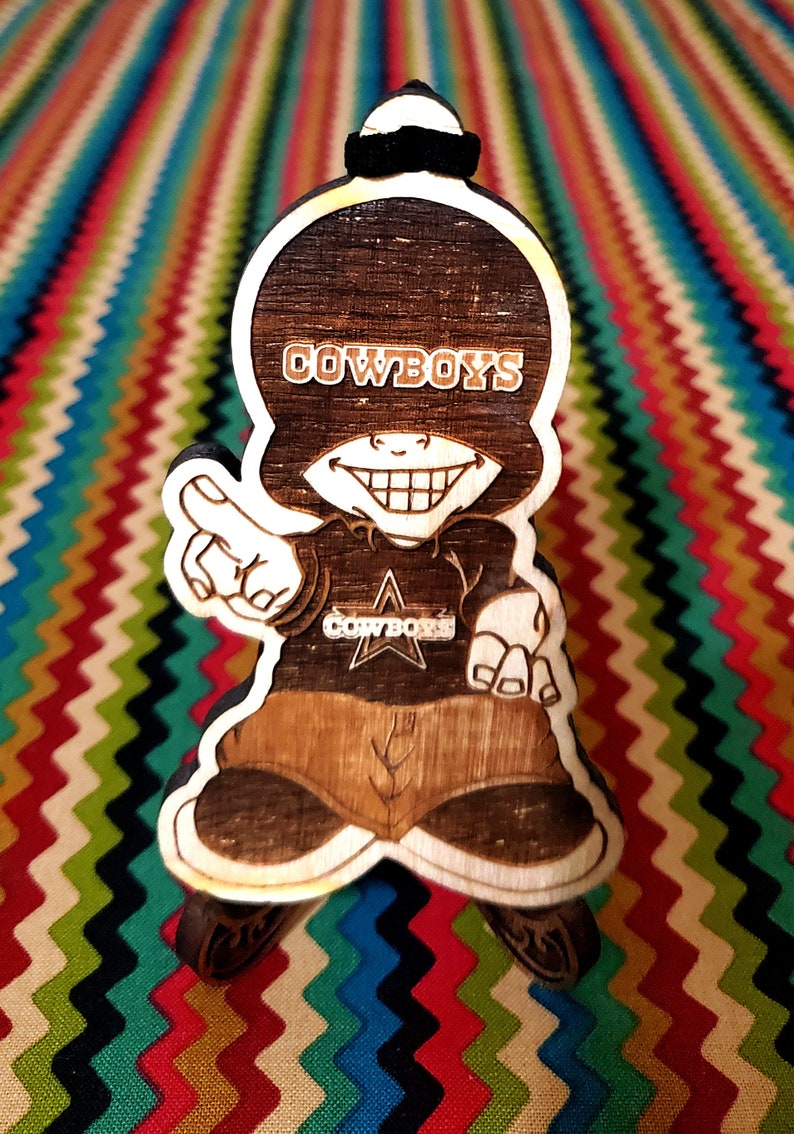 May include: A wooden figurine of a cartoon character wearing a hat and a shirt with the word "COWBOYS" on it. The character is smiling and has a star on its shirt.