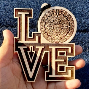 Könnte beinhalten: Holzornament mit der Aufschrift "LOVE" und einem dekorativen aztekischen Kalendermotiv. Die Buchstaben sind in einem kräftigen Blockstil mit kontrastierendem Holzmaserung gehalten. Ein schwarzes Band ist zum Aufhängen befestigt.