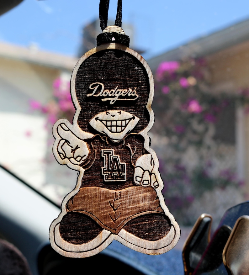 May include: A wooden car charm featuring a cartoon character with a mischievous grin, wearing a baseball cap that says "Dodgers" and a jersey with the LA logo. The charm is brown and has a wood grain texture.