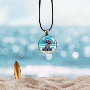 Op de afbeelding: Een ronde hangerketting met een stripfiguur aan een zwart koord. De hanger heeft een blauw-witte achtergrond met een stripfiguur. Een houten surfplank staat in het zand op de achtergrond.