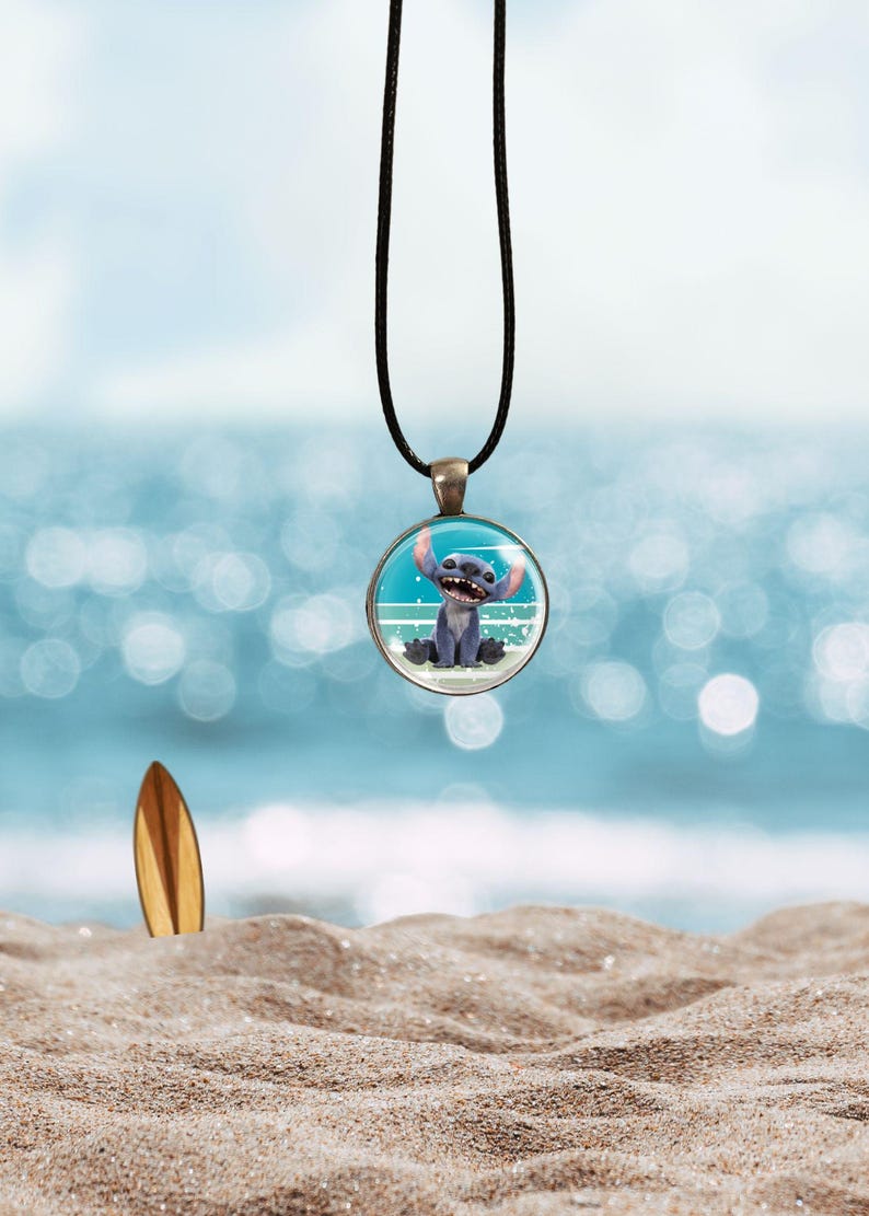 Op de afbeelding: Een ronde hangerketting met een stripfiguur aan een zwart koord. De hanger heeft een blauw-witte achtergrond met een stripfiguur. Een houten surfplank staat in het zand op de achtergrond.