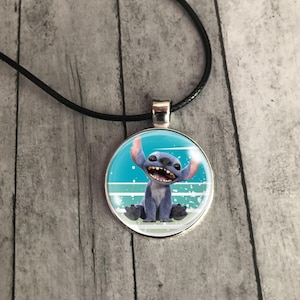 Op de afbeelding: Een ronde hangerketting met een cartoonafbeelding van Stitch uit Lilo & Stitch. De hanger heeft een zilverkleurige rand en hangt aan een zwart koord. De achtergrond van de hanger is blauw en wit.