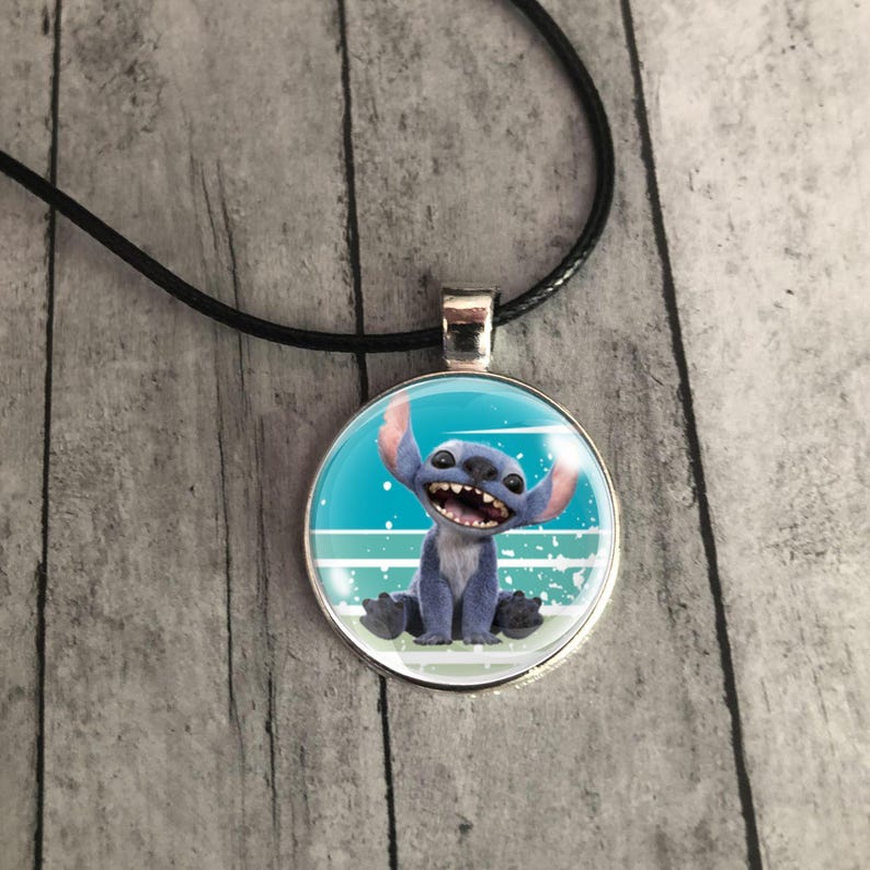 Op de afbeelding: Een ronde hangerketting met een cartoonafbeelding van Stitch uit Lilo & Stitch. De hanger heeft een zilverkleurige rand en hangt aan een zwart koord. De achtergrond van de hanger is blauw en wit.