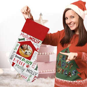 May include: White Christmas stocking with a red cuff and a personalized dog house design. The stocking features a photo of a woman and her dog, along with the name "Molly" repeated throughout the design. The stocking is held by a woman wearing a red sweater with a green Christmas tree design.