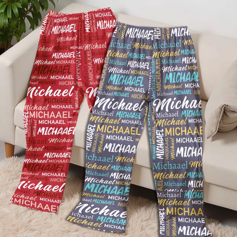 May include: Three pairs of pajama pants with the name "Michael" printed repeatedly in various colors. One pair is red, another is gray, and the third is a darker gray. The pajamas are displayed on a white couch.
