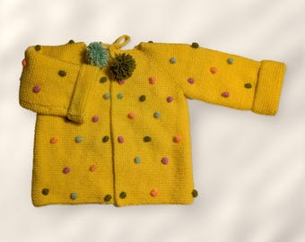 May include: A bright yellow knit cardigan featuring long sleeves with folded cuffs and a round neckline with a tie closure. The garment is decorated with small, multi-colored pom-poms in shades of pink, blue, orange, and olive green.