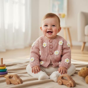 Puede incluir: Un bebé sonriente con un cárdigan rosa tejido adornado con margaritas blancas. El bebé está sentado sobre una manta beige, rodeado de juguetes de madera, incluyendo una torre apilable y un coche. También hay un oso de peluche.