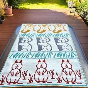 May include: A handmade crochet blanket featuring a repeating pattern of koalas and other Australian animals. The blanket has a white background with accents of gray, teal, orange, and yellow. The blanket is rectangular.