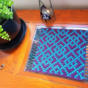 Puede incluir: Un posavasos cuadrado de ganchillo de color turquesa y morado con flecos en todos los lados. El posavasos está sobre una mesa de madera con tapa de cristal.