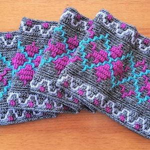 May include: A gray crocheted scarf with a geometric pattern in pink, teal, and white. The scarf has fringe on both ends.