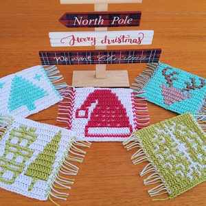 Puede incluir: Un conjunto de cinco posavasos cuadrados hechos a mano, de ganchillo, con bordes con flecos. Cada posavasos presenta un diseño navideño diferente, incluyendo un árbol de Navidad, un gorro de Papá Noel, un reno y patrones de copos de nieve. Un letrero de madera con "North Pole" y "Merry Christmas" está en el fondo.