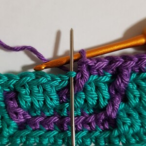May include: Close-up of a crochet project in progress. A teal and purple yarn is being worked with a silver crochet hook. The hook is inserted into a stitch on the teal yarn.
