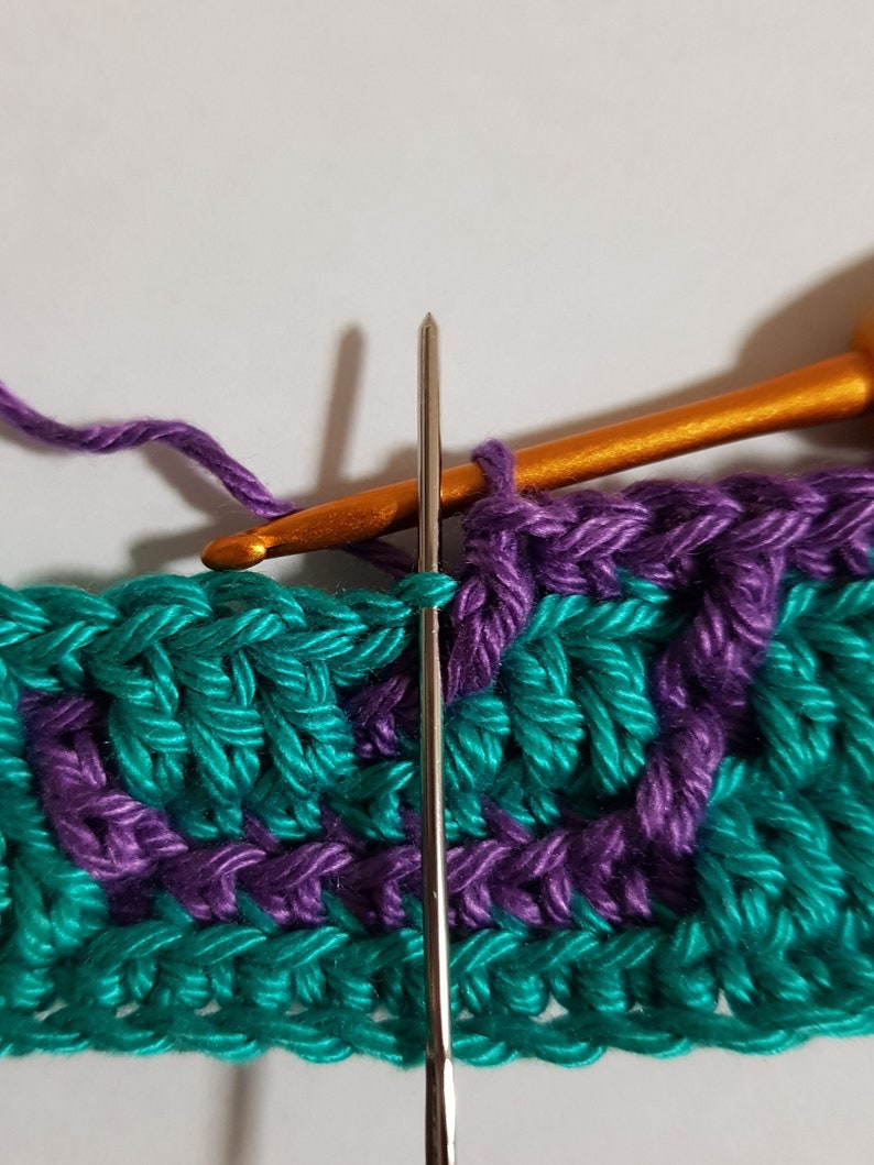 May include: Close-up of a crochet project in progress. A teal and purple yarn is being worked with a silver crochet hook. The hook is inserted into a stitch on the teal yarn.